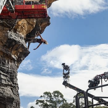 A scene from the movie 'Apex' featuring a stunt performer hanging from a rocky cliff with a film crew and equipment visible in the foreground. Bright blue sky and clouds provide the backdrop.