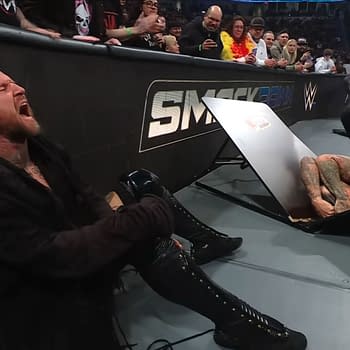 Aleister Black sits on the floor, visibly in pain, next to a Slim Jim-themed table that is broken and positioned upside down, with Damian Priest lying on the ground beneath it, during a WWE SmackDown event.