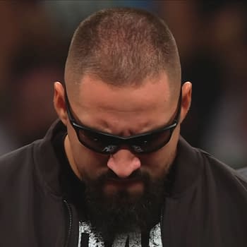 Bron Breakker, wearing sunglasses and a dark jacket, appears focused and prepared for an upcoming confrontation, with his head down and a serious expression. Paul Heyman can be seen in the background, watching him intently.