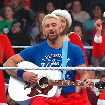 Joe Hendry, wearing a blue t-shirt, sings while holding an acoustic guitar, surrounded by women in Santa Claus outfits. The scene takes place inside a wrestling ring, with fans visible in the background.