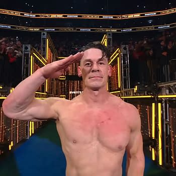 John Cena salutes the fans with a serious expression while shirtless, standing on a brightly lit stage following his last match at Saturday Night's Main Event. A crowd is visible in the background, cheering him on.