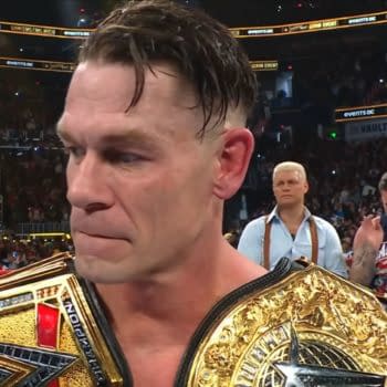 John Cena, holding two championship belts, expresses emotion as he stands amidst a cheering audience following his final match at Saturday Night's Main Event. Peers in the background show their support while the crowd is visibly excited.