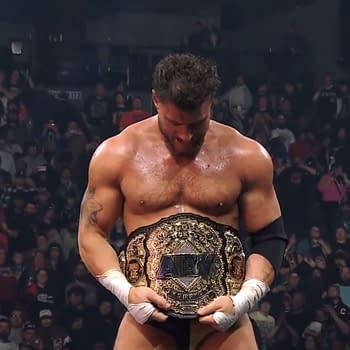 A wrestler stands in the ring, holding the AEW World Championship belt and looking down at it, surrounded by an excited crowd of fans. The atmosphere is electric, reflecting the significance of the moment.