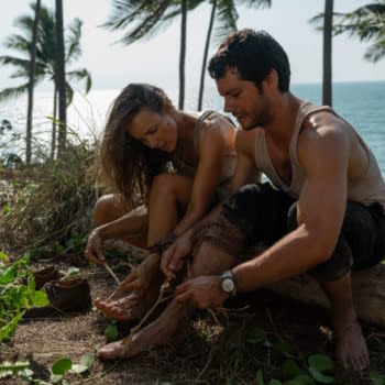 In a scene from 20th Century Studios' 'SEND HELP,' characters Linda Liddle and Bradley Preston work together on a beach, tying makeshift bindings on their feet. The backdrop features a serene ocean view and palm trees.