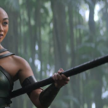 A female character, dressed in a green and gold warrior outfit, stands confidently in a lush forest environment, holding a long spear. The sunlight filters through the trees, creating a mystical atmosphere.