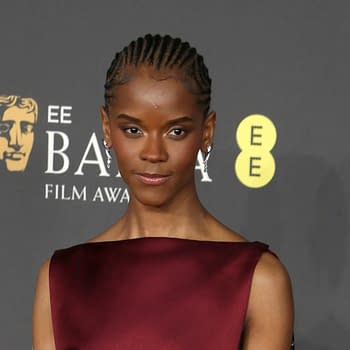 Letitia Wright poses confidently at the BAFTA Film Awards, wearing a stylish maroon dress and statement earrings, with the BAFTA logo visible in the background.