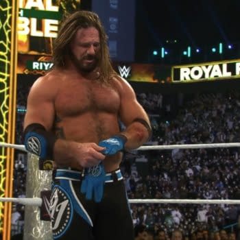 A wrestler with long hair, wearing a blue and black outfit, is taking off his blue gloves inside a wrestling ring after losing a retirement match. The crowd is visible in the background, with dim lighting and a large 'Royal Rumble' sign overhead.