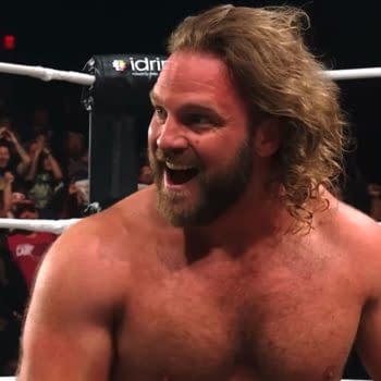 Hangman Adam Page celebrates a victory in the wrestling ring, visibly excited with a wide smile, while fans in the background cheer enthusiastically.