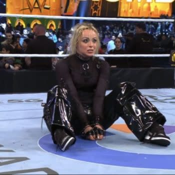 A female wrestler in a black bodysuit, styled with glossy pants, sits on the wrestling ring after winning the Women's Rumble match. The crowd in the background shows mixed expressions of interest.