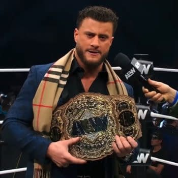 A wrestler holds the AEW World Championship belt while being interviewed by another individual, dressed in a blazer and a plaid scarf. The wrestling ring is visible in the background with an audience.