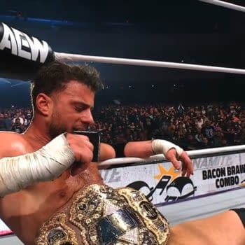 A visibly exhausted wrestler rests in the ring after a title defense at AEW Dynamite, gripping his championship belt while seated on the mat. The large crowd is visible in the background, capturing the atmosphere of the event.