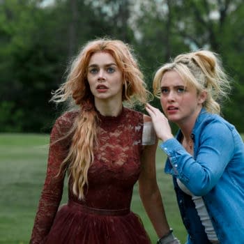 A scene from the film 'Ready or Not 2: Here I Come', featuring two women: one with long, wavy red hair wearing a burgundy lace dress, and the other with blond hair in a ponytail, dressed in a blue denim shirt. They appear anxious and are looking off-screen, set against a green outdoor background.