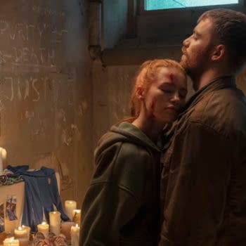 A tense scene from The Strangers - Chapter 3, featuring characters Maya and Gregory in an emotionally charged embrace amidst a dimly lit background adorned with candles and photographs. The setting conveys a sense of desperation and mystery, with cryptic words scrawled on the walls.