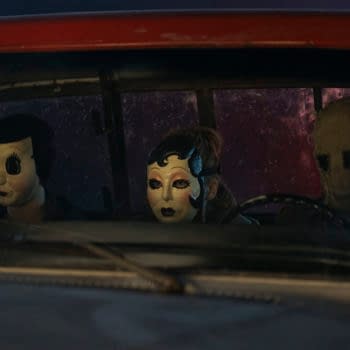 Three figures wearing creepy masks sit in a truck's front seat, creating a tense atmosphere. This still is from 'The Strangers - Chapter 3,' with a backdrop featuring ominous colors.
