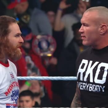 Sami Zayn and Randy Orton face each other in the wrestling ring during an episode of WWE SmackDown, with an enthusiastic crowd in the background. Zayn wears a white shirt, and Orton is in a black t-shirt with 'RKO' printed on it.