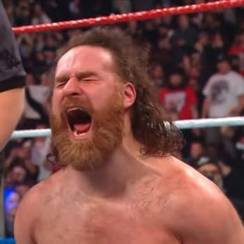 Sami Zayn, with long hair and a beard, celebrates a victory in a wrestling ring, expressing intense emotion with a loud shout. A referee is visible by his side, and a cheering crowd can be seen in the background.