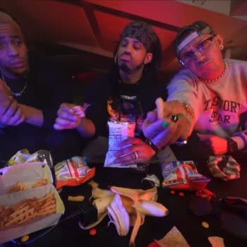 Three members of The Rascalz relax backstage on AEW Collision, seated around a table filled with snacks, including fries and various chips. The dimly lit setting features colorful ambient lighting, highlighting their expressions as they interact with each other.