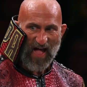 A bald, bearded wrestler wearing a red and black outfit with a high collar has a fierce expression on his face. His blue eyes are wide open, conveying intensity.