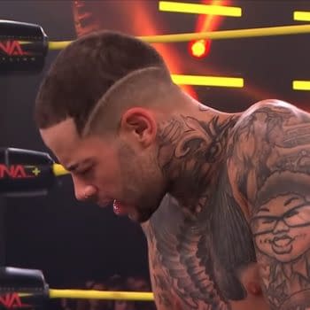 A wrestler with intricate tattoos on his bare torso, looking down, in a wrestling ring with yellow ropes and bright lights around. The wrestler has a unique haircut with a shaved line on the side of his head.
