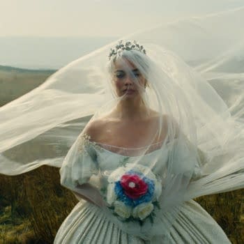 A bride in a flowing white gown stands in a grassy field, her veil billowing in the wind. She holds a bouquet of colorful flowers, conveying a sense of wistfulness and beauty.