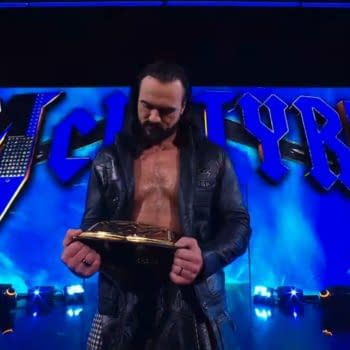 Drew McIntyre stands on stage, holding his championship belt and looking down at it. The background features blue lights and the name 'McIntyre' displayed prominently.