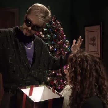 A man in sunglasses and a patterned black jacket gestures dramatically while holding an open gift box, with a Christmas tree lit up in the background. An intrigued woman with curly hair watches.
