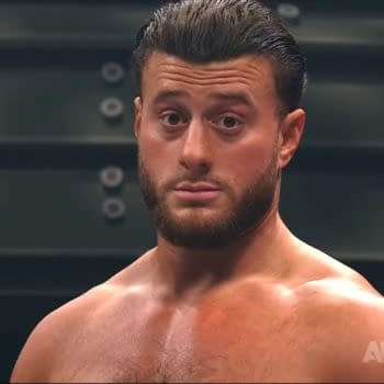 A professional wrestler shows a serious expression in response to a loud crowd chant during a wrestling match. The background features blurred steel structures typical of a wrestling arena.