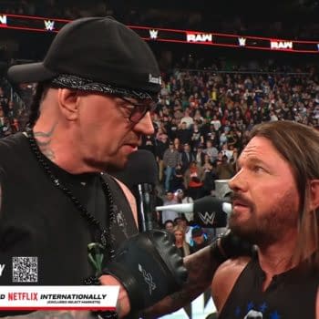 The Undertaker, wearing a black bandana and glasses, speaks into a microphone, announcing his WWE Hall of Fame induction to AJ Styles, who looks surprised. The audience is visible in the background, filling the arena with anticipation.