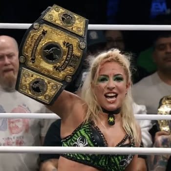 A female wrestler joyfully holds up the AEW Women's World Championship belt, wearing a green and black outfit. The audience is visible in the background, with various expressions of excitement.