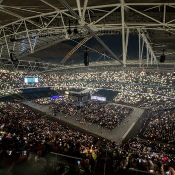 A wide shot of Wembley Stadium during the AEW All In event, filled with fans holding up lights, creating a sparkling effect in the arena. The atmosphere is electric with a large wrestling ring at the center.
