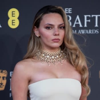 A woman with long, wavy hair wearing a glamorous pearl necklace and a strapless white gown poses confidently at the BAFTA Film Awards backdrop.