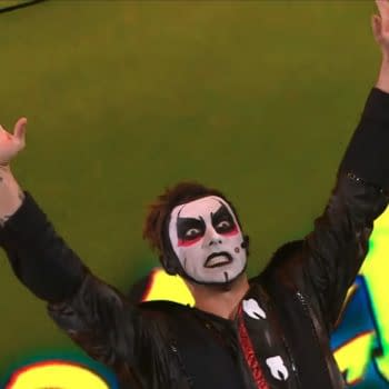 A performer in a dramatic costume, featuring bold face paint and a black jacket, raises both hands in the air during a WWE Raw event. The performer has a striking facial expression that adds to the comedic effect.