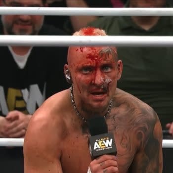 Darby Allin, showing signs of a brutal match with blood on his face and a determined expression, holds a microphone while addressing the audience on AEW Dynamite. He has visible tattoos, a chain around his neck, and a mohawk hairstyle.