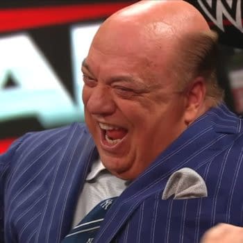 Paul Heyman is laughing joyfully while seated at a WWE Raw event, wearing a pinstriped suit and a tie. The background features WWE branding, creating an energetic atmosphere.
