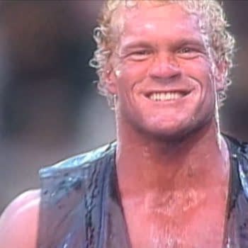 Sycho Sid Eudy, a wrestler with curly blonde hair and a wide smile, wearing a black vest, stands confidently in front of a blurred background. The wrestler appears sweaty and is showcasing a victorious expression.