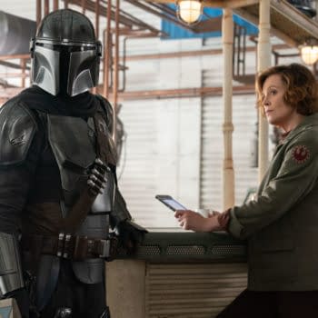 Mandalorian character in full armor stands next to Colonel Ward, who is seated at a bar-like counter, both appearing to engage in conversation. This scene is from Lucasfilm's 'The Mandalorian and Grogu.'