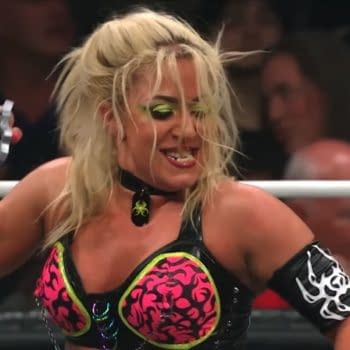 A female wrestler, Thekla, is in the ring during a match on AEW Dynamite, raising her fist with brass knuckles visible. She has long blonde hair, dramatic makeup, and wears a colorful outfit with a choker necklace.