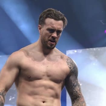 A muscular wrestler with tattoos stands shirtless in a wrestling arena, appearing intense and focused under dramatic lighting. The background features a large screen and beams of light, enhancing the electrifying atmosphere.