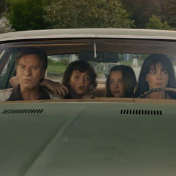 A dramatic scene in a vintage car featuring a man and three children, each displaying varied expressions of concern and intensity while looking out the windshield. The background includes a suburban setting with green trees and houses.