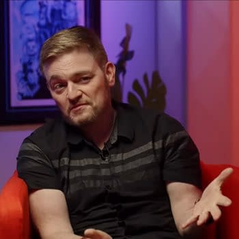 Scott Snyder gives a DC Comics interview. In the background, there are colorful lights and a framed artwork, along with a Batman figurine on a table nearby.