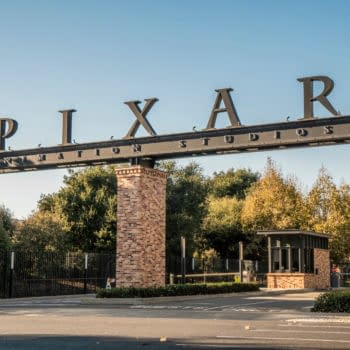 The entrance gate to Pixar Animation Studios, featuring large black lettering that spells "PIXAR" atop a brick structure, set against a clear blue sky and green trees.