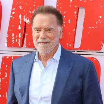 Arnold Schwarzenegger poses at a red carpet event, wearing a blue suit and light shirt, with bold red promotional signage in the background.