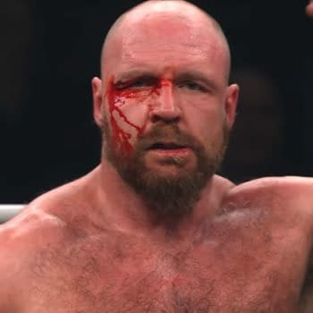 A muscular man with a bald head and beard displays a bloodied face, raising his arm triumphantly after a wrestling match. The marks of his battle are evident on his body.