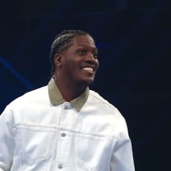 Lil Yachty smiles under blue stage lights on WWE SmackDown, wearing a white jacket.