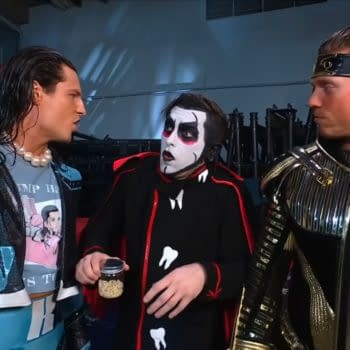 In a backstage scene from WWE SmackDown, Kit Wilson, dressed in shimmering blue attire, appears to reject a proposal from Danhausen, who is in a black costume with white face paint, holding a jar of snacks. Another wrestler, also dressed in metallic gold, observes the interaction.
