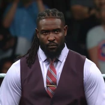 A man with long braided hair wearing a dark vest and light purple shirt stands confidently in a wrestling ring, appearing serious against a cheering crowd in the background. He is dressed for a formal occasion, enhancing his commanding presence.