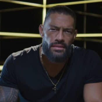 Roman Reigns, a professional wrestler, sits in front of an illuminated backdrop, looking directly at the viewer with an intense expression.
