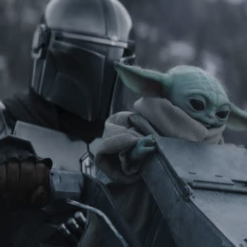 The Mandalorian, wearing a silver helmet and armor, holds onto a vehicle while Grogu, a small green creature with large ears, sits beside him wrapped in a grey robe. The scene conveys a moment of adventure in a snowy landscape.