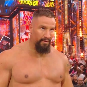 A muscular man with a beard stands in a wrestling ring, focused on the action, surrounded by an excited crowd and colorful lighting from the event's backdrop.