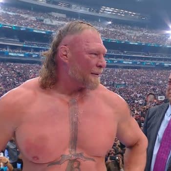 A muscular wrestler with long hair and a visible chest tattoo stands in a wrestling ring, looking pensive amidst a large crowd. Another man in a suit is standing next to him, appearing emotional.